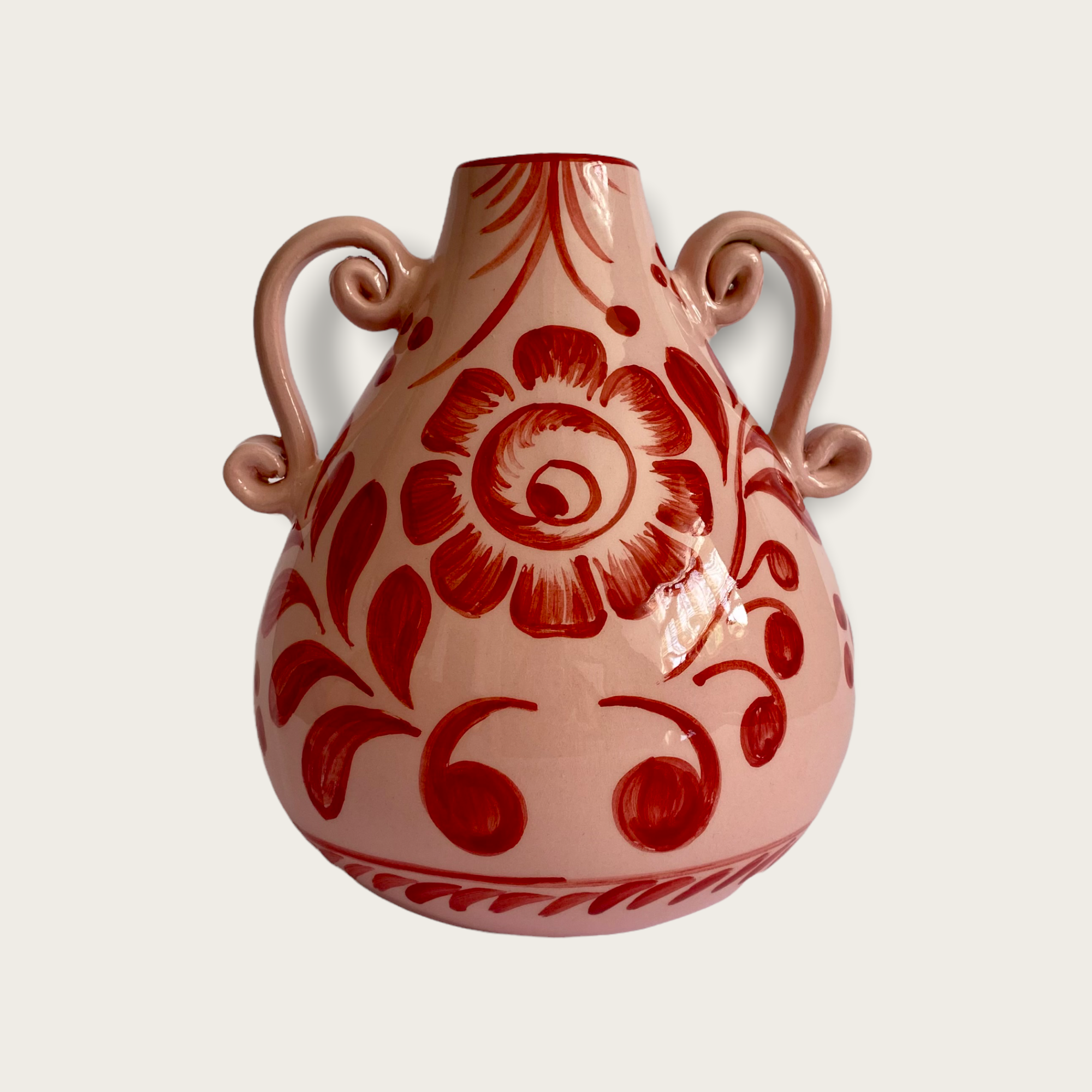 A pink vase with a red floral pattern hand painted. The vase is a sculptured shaped with two ornate handles.