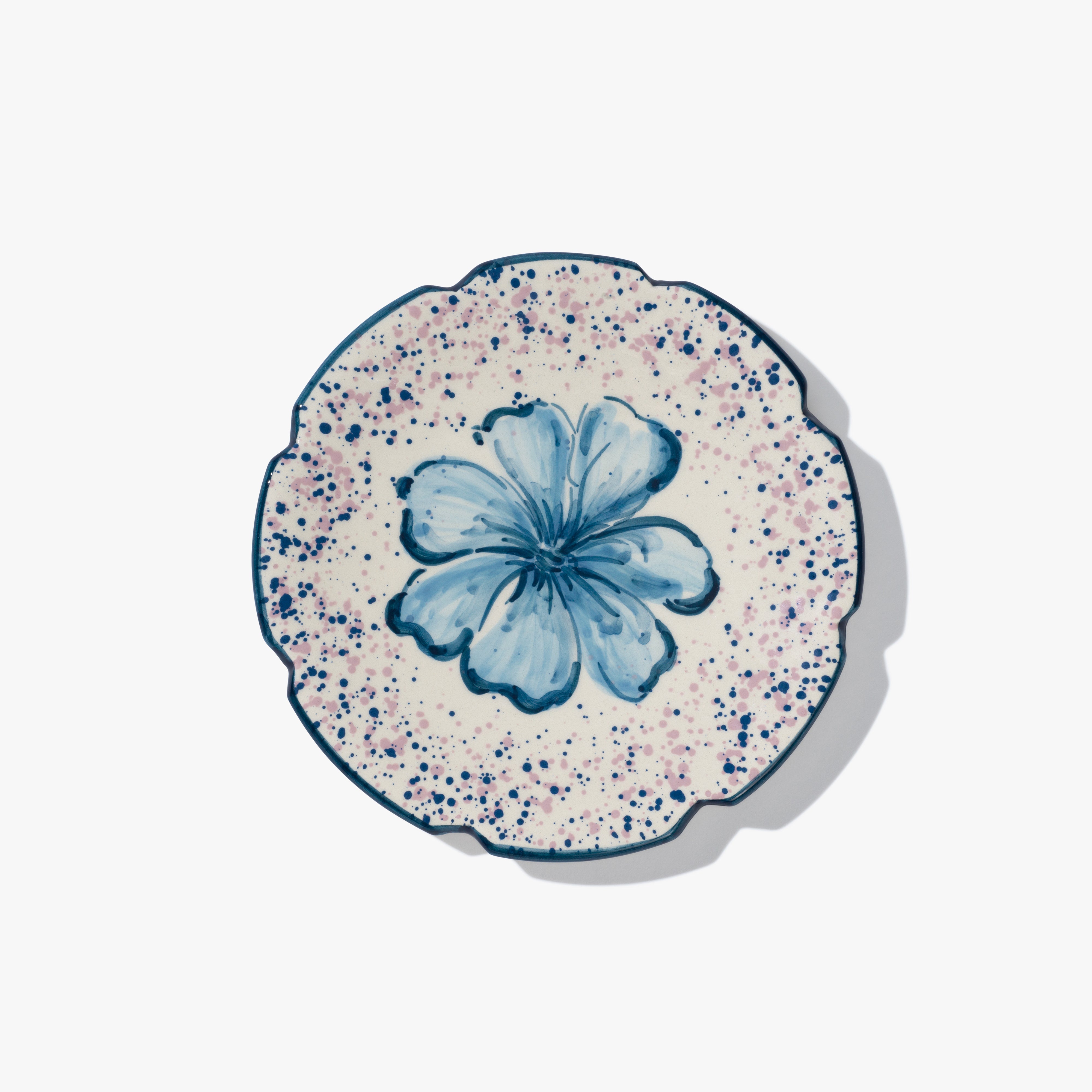 A round ceramic plate with a blue flower in the centre. The plate has a scalloped edge, which is also painted blue and and blue and pink speckled patten around the flower.