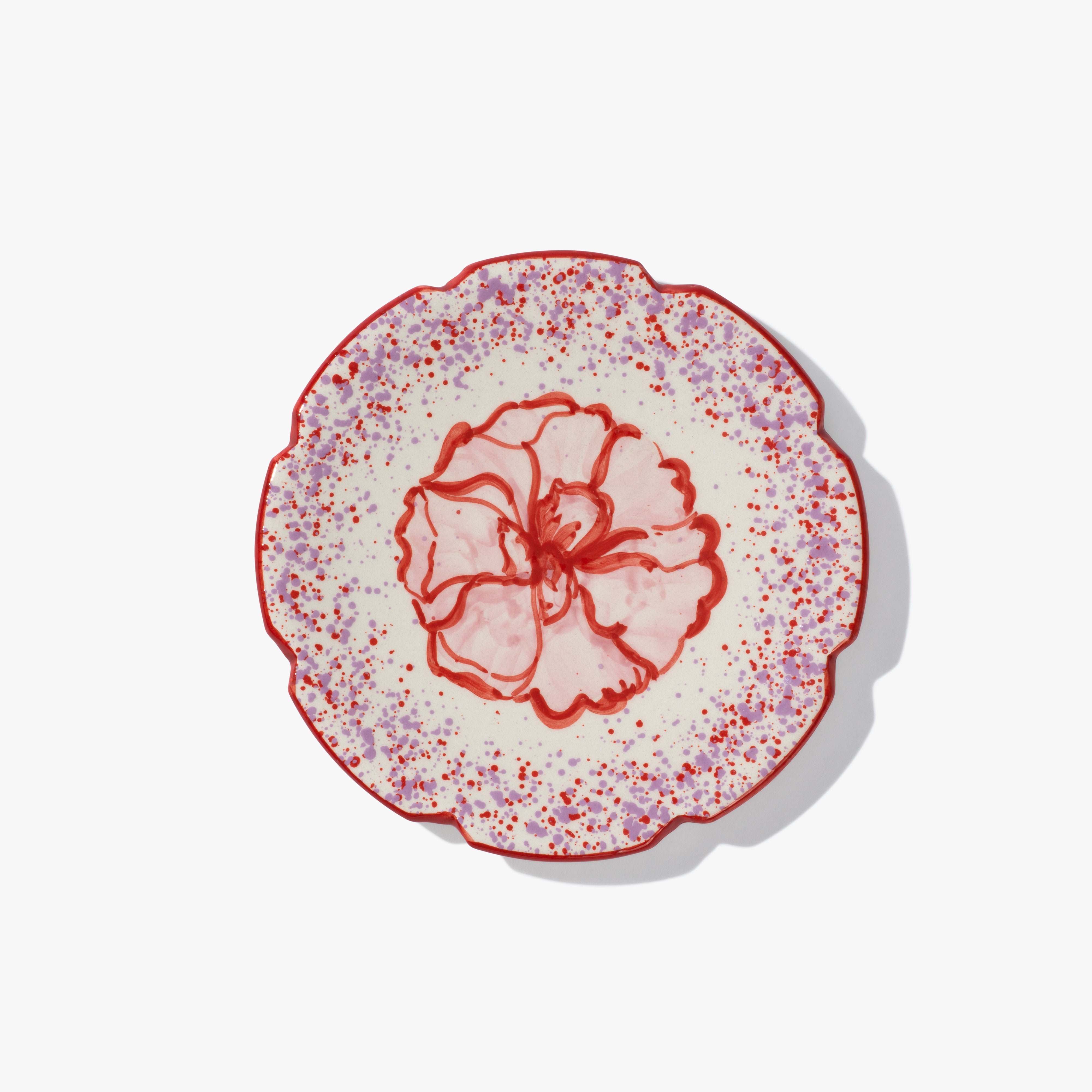 A round ceramic plate with a pink and red flower in the centre. The plate has a scalloped edge, which is also painted red and and red and lilac speckled patten around the flower.