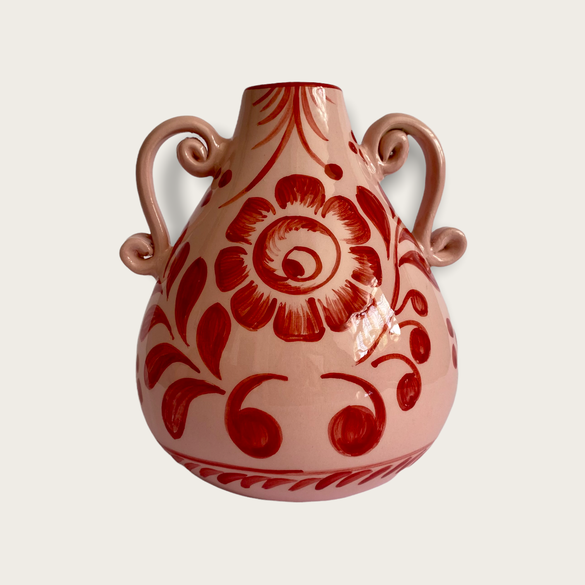 A pink vase with a red floral pattern hand painted. The vase is a sculptured shaped with two ornate handles. 