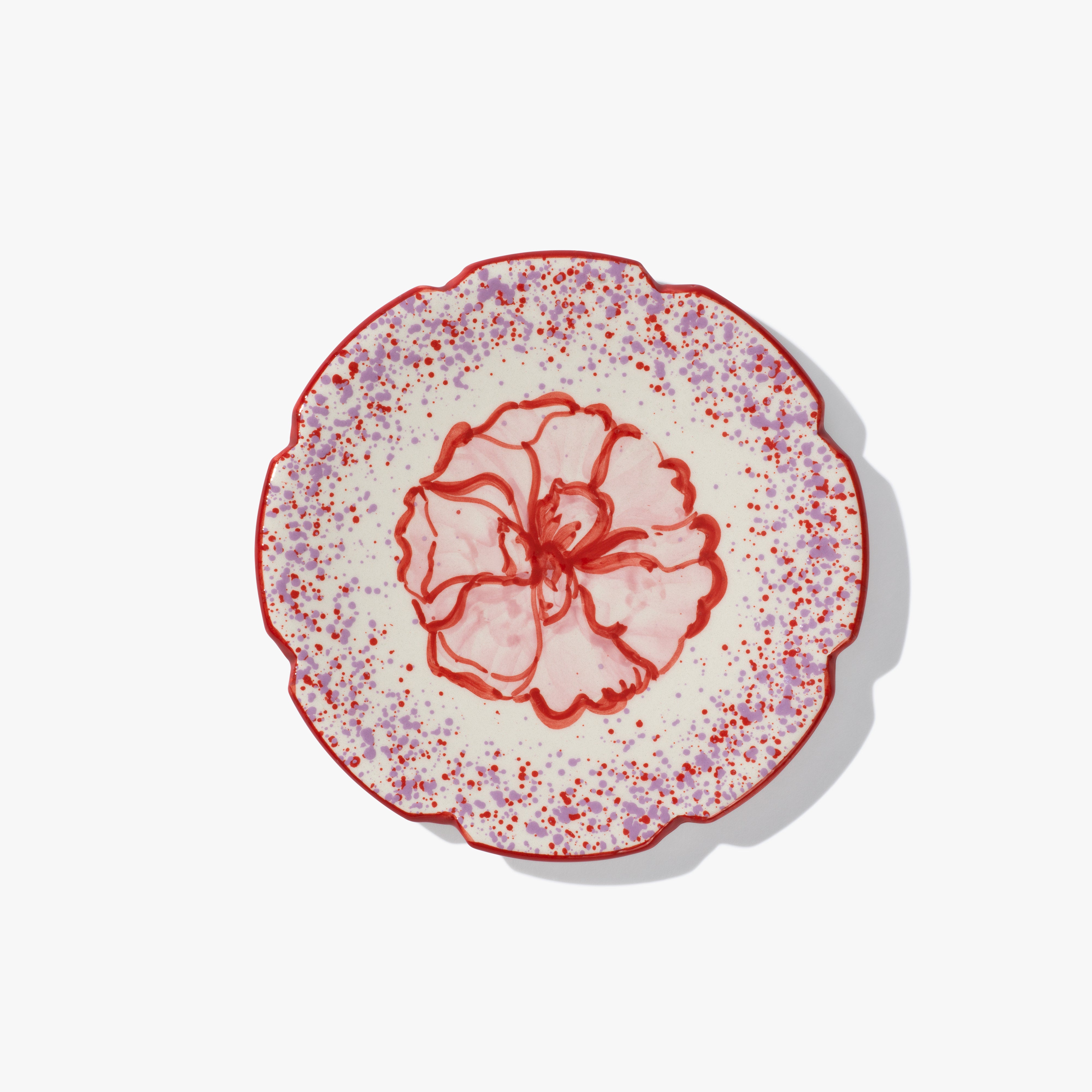 A round ceramic plate with a pink and red flower in the centre. The plate has a scalloped edge, which is also painted red and and red and lilac speckled patten around the flower. 