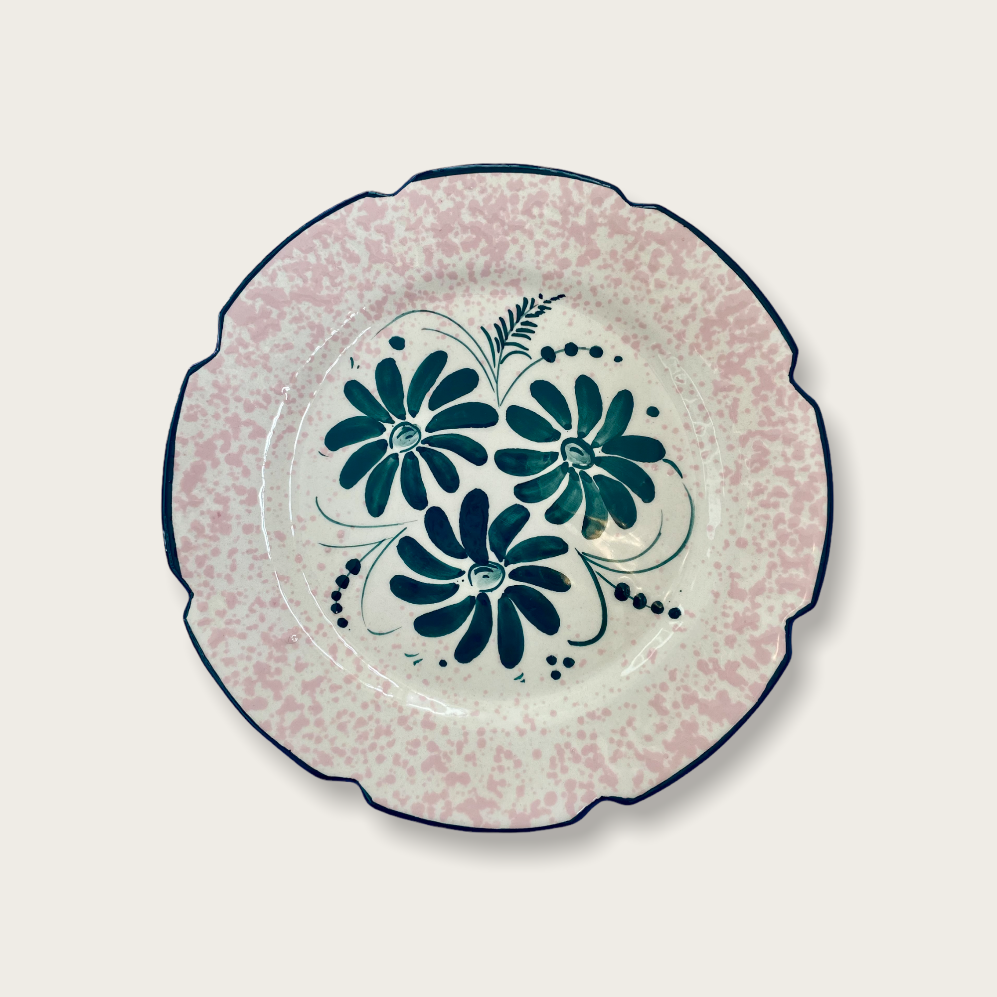 A vaisselle leonie plate which has a scalloped edge, rimmed in deep green and complimantery forest green flowers in the centre, surrounded by a light pink speckle pattern on the rim. 
