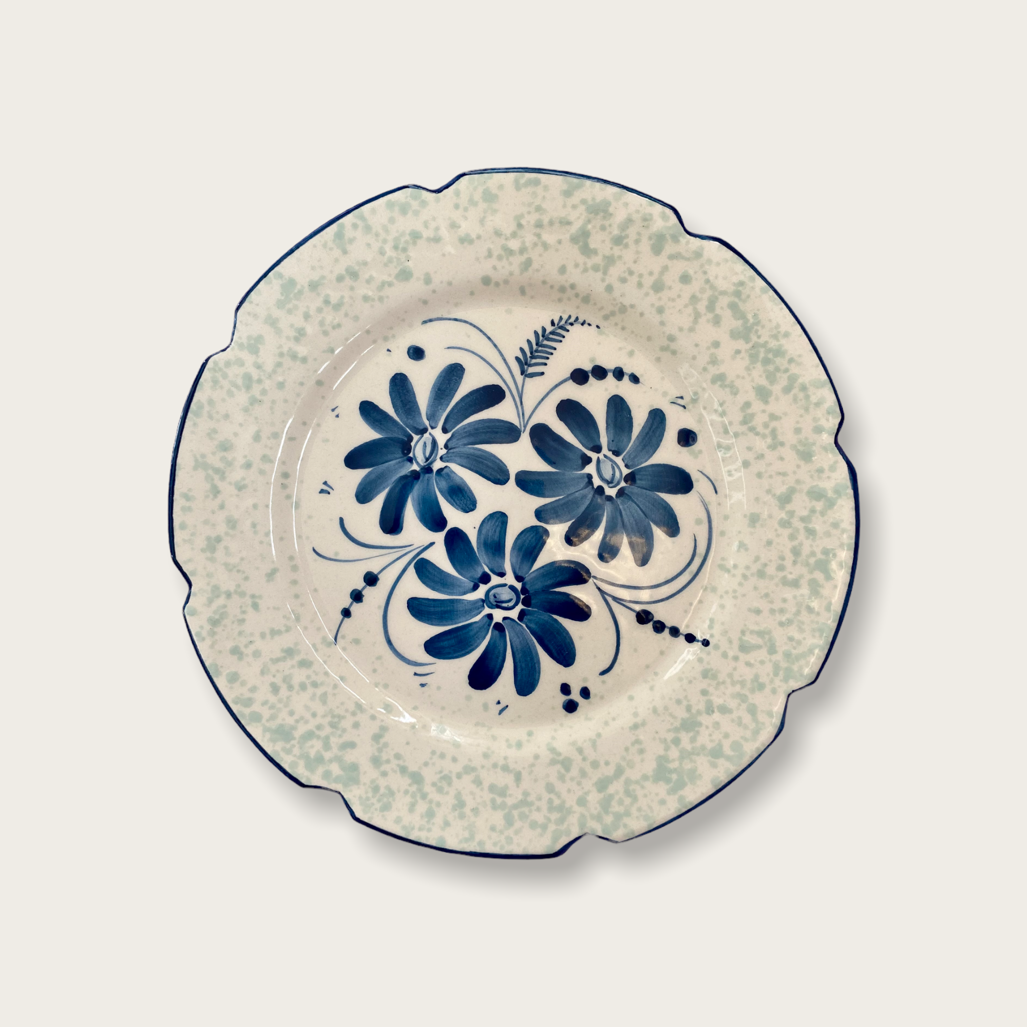 Aceramic plate that features a trio of blue/ indigo flowers and an indigo scalloped edge. The floral design is surrounded by a contrasting and unique scalloped rim with a mint green speckled pattern. .